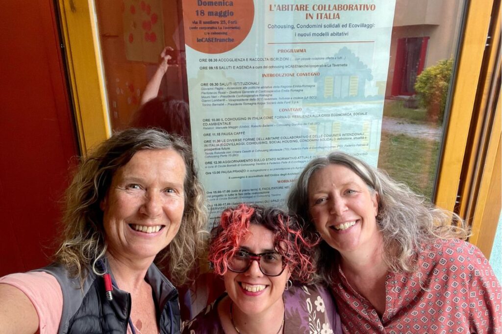 Tre partecipanti al convegno sull’abitare collaborativo al cohousing Case Franche davanti alla locandina ufficiale dell’evento.