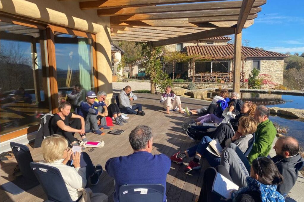 Partecipanti del Capodanno detox ad Autosufficienza seduti in cerchio sotto il pergolato durante una sessione di gruppo all’aperto, con il panorama dell’Appennino romagnolo sullo sfondo.