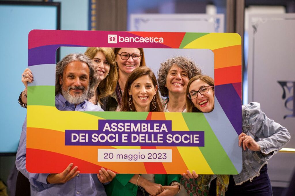 Gruppo di soci e socie di Banca Etica sorride dietro una cornice arcobaleno con la scritta assemblea dei soci e delle socie 20 maggio 2023
