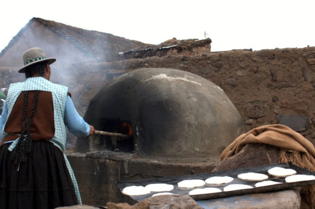 Donna in abiti tradizionali boliviani che inforna il pane in un forno di terra in un villaggio andino, durante un viaggio di turismo responsabile.